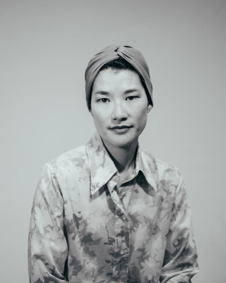 Black and white medium shot of an Asian woman wearing a floral shirt and head wrap.