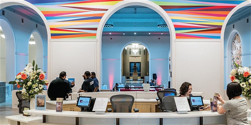 This is a photo of Englehard Court at the Newark Museum. A female museum worker assists a visitor at the admissions desk.