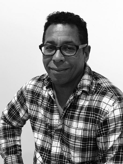 Black and white medium shot of a black man smiling and wearing a plaid shirt.