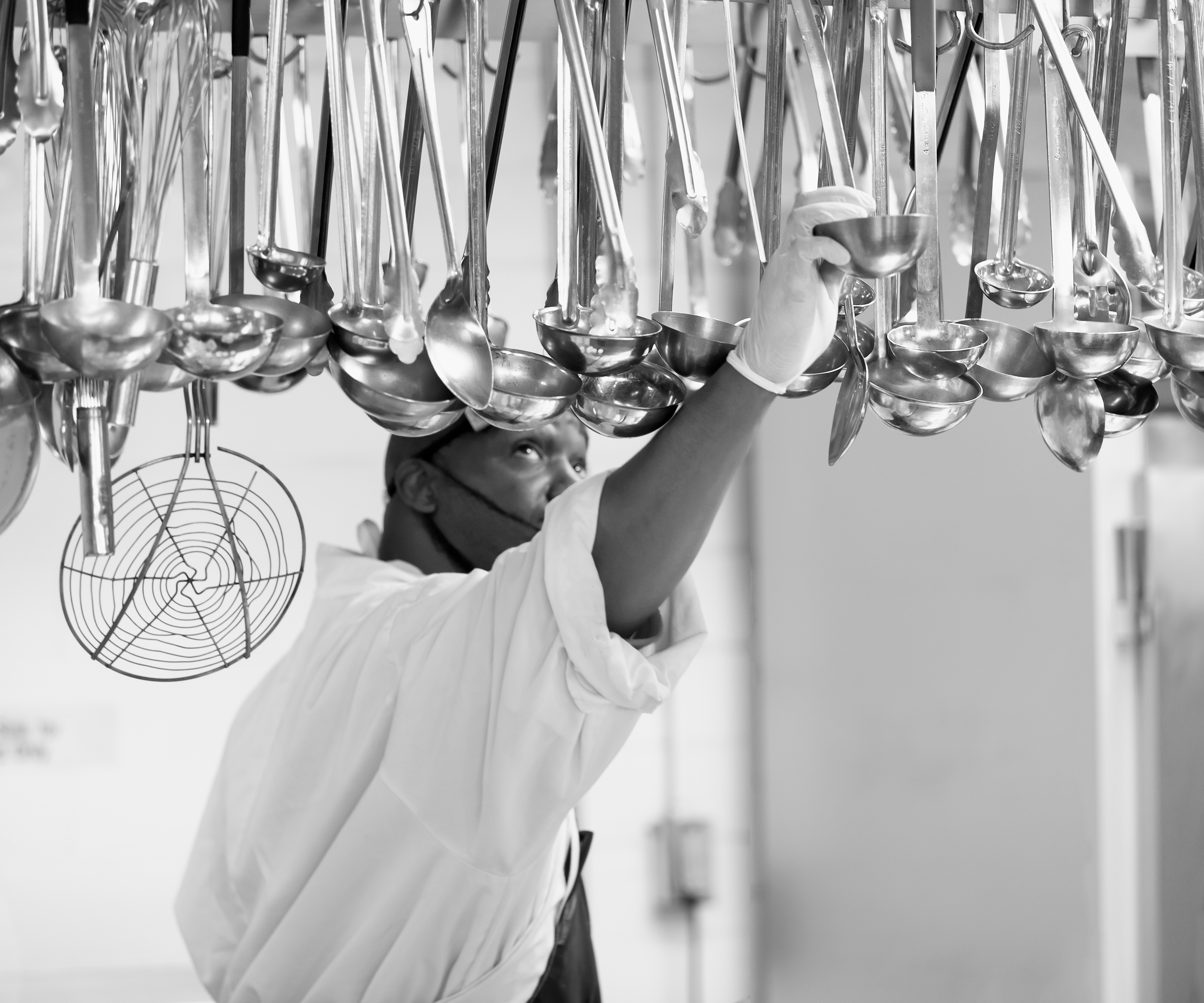 A man in chefs whites reaches up to grab a ladle hung from a rack above his head