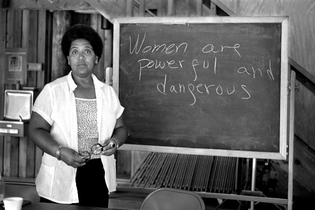 A Black woman stands next to a chalk board that reads, "Women are powerful and dangerous."