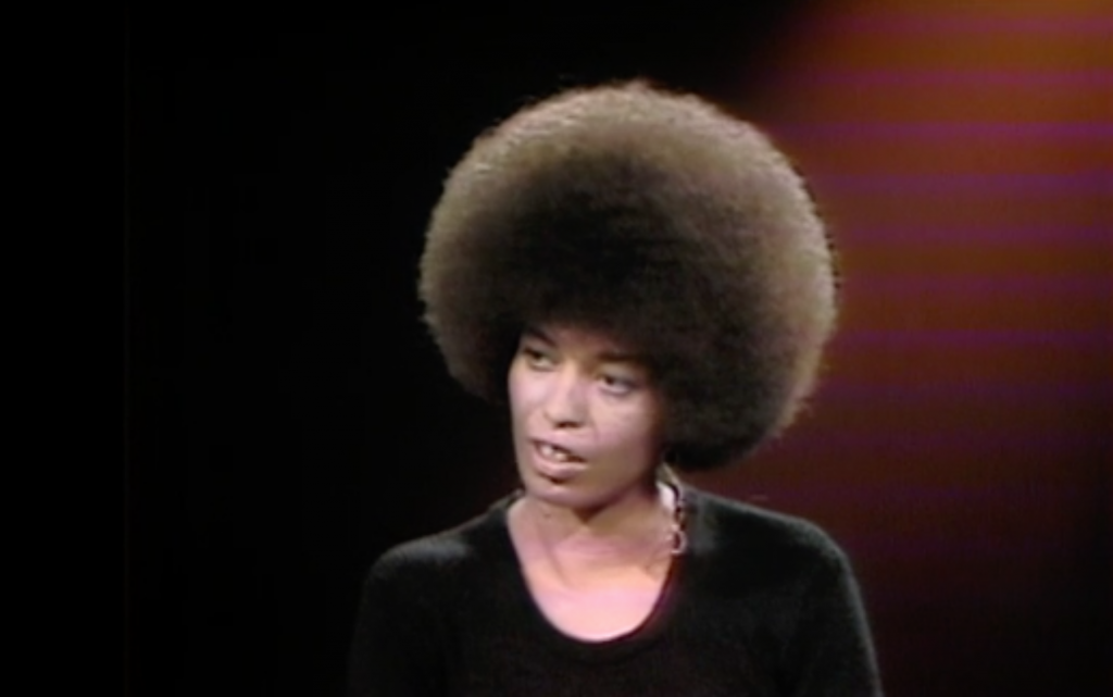 A medium shot of a Black woman with an afro speaking against a black and red background